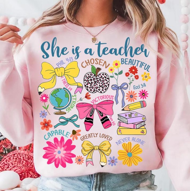 以下が含まれることがあります： 「She is a teacher」という文字と、リボン、花、本、地球儀のデザインが施されたピンク色のスウェットシャツ。デザインには、聖書の言葉や「Chosen」、「Beautiful」、「Victorious」、「Capable」、「Greatly Loved」、「Never Alone」などの言葉も含まれています。