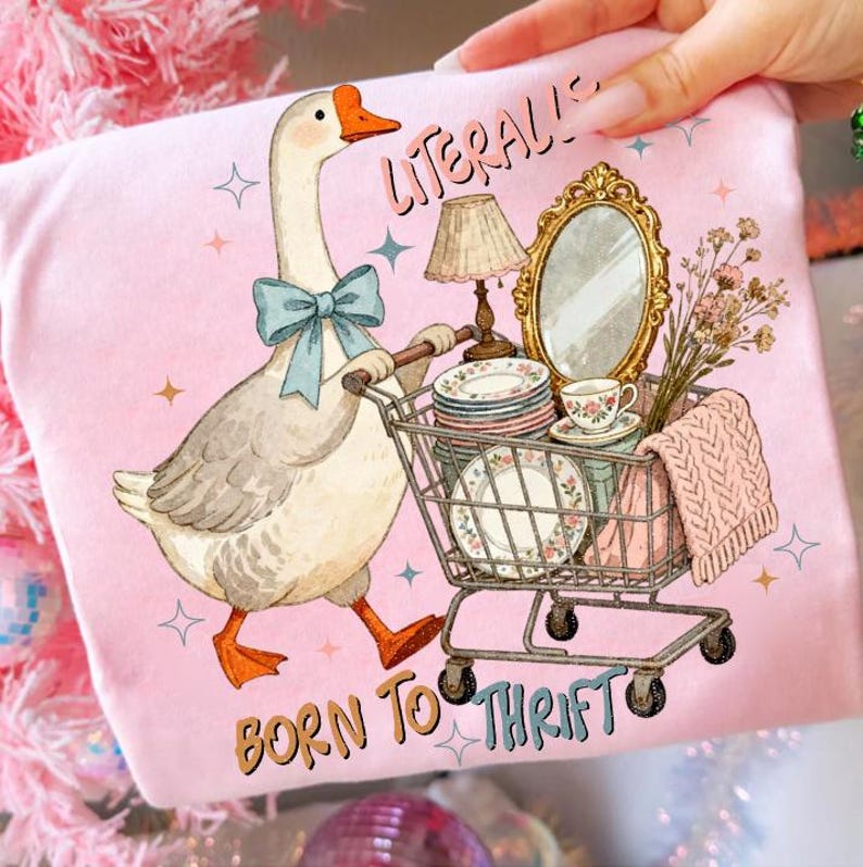 May include: Pink t-shirt featuring a whimsical illustration of a goose pushing a shopping cart filled with vintage items. The goose wears a blue bow tie. Text on the shirt reads "Literally Born to Thrift."