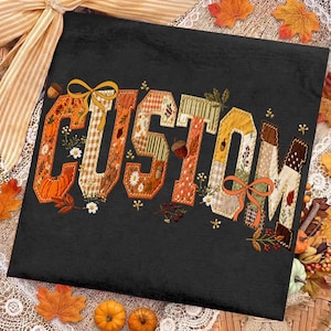 May include: A black t-shirt featuring the word CUSTOM in large, block-style letters. Each letter is filled with a patchwork pattern of autumn-themed motifs, including pumpkins, acorns, fall leaves, flowers, and plaid designs in orange and brown.