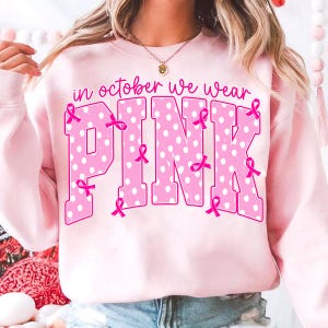 Puede incluir: Una sudadera de cuello redondo rosa claro con el texto "in october we wear PINK" en una fuente de letras grandes y negritas. Las letras est&aacute;n rellenas con un patr&oacute;n de lunares blancos y decoradas con peque&ntilde;os lazos rosas de concienciaci&oacute;n.