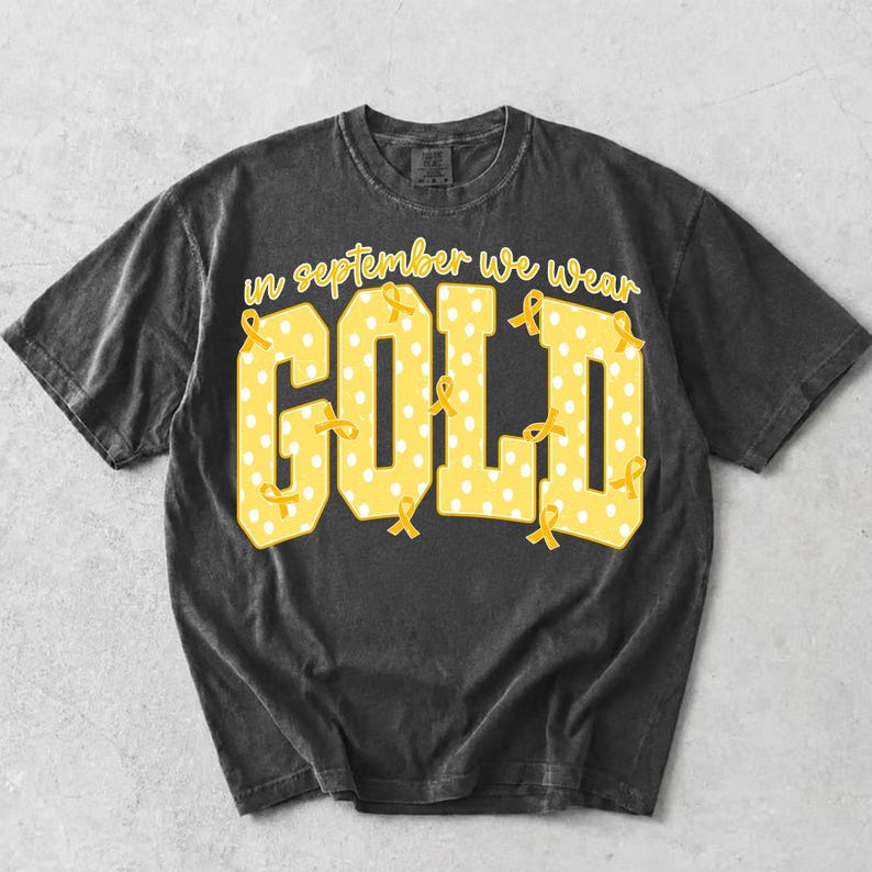 May include: A charcoal grey short-sleeve t-shirt featuring the yellow text "in september we wear GOLD" in a block font. The letters are filled with a white polka dot pattern and decorated with small yellow awareness ribbons throughout the design.