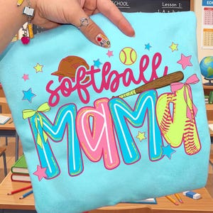 May include: Light blue sweatshirt with the words "softball MAMA" in pink and blue lettering. The design includes a softball, baseball bat, and stars. A brown baseball cap and a softball glove are also featured in the design.