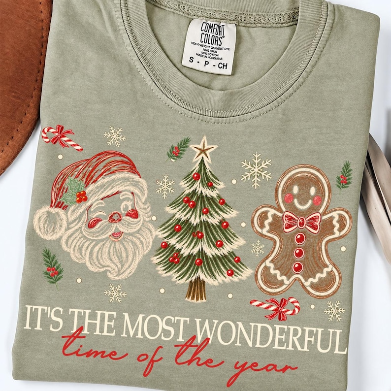 K&ouml;nnte beinhalten: Ein salbeigr&uuml;nes T-Shirt mit einem Grafikdruck von Weihnachtsmann, einem geschm&uuml;ckten Weihnachtsbaum und einem Lebkuchenmann. Der Text lautet "IT'S THE MOST WONDERFUL time of the year". Das T-Shirt hat ein Comfort Colors Markenetikett am Hals.