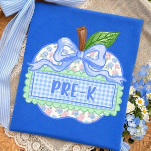 Op de afbeelding: Een koningsblauw t-shirt met een centrale appelvormige afbeelding. De appel heeft een bloemenpatroon, een lichtblauwe, geruite rechthoekige banner met de tekst PRE-K, een lichtblauwe strik, een bruine steel en een groen blad met een schulprand.