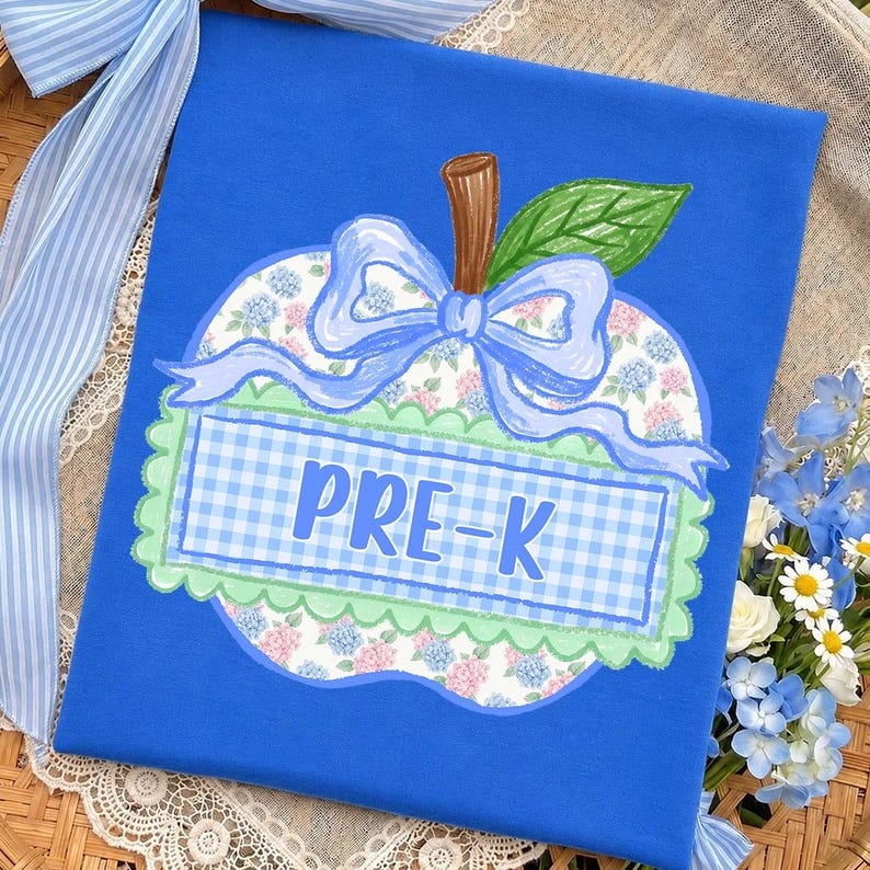Op de afbeelding: Een koningsblauw t-shirt met een centrale appelvormige afbeelding. De appel heeft een bloemenpatroon, een lichtblauwe, geruite rechthoekige banner met de tekst PRE-K, een lichtblauwe strik, een bruine steel en een groen blad met een schulprand.
