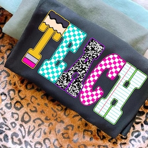 May include: Dark gray t-shirt with the word "TEACH" in colorful, block letter designs. The letters feature patterns like checkered, notebook paper, and a pencil. The shirt is folded on top of other shirts.