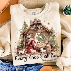 May include: Cream-colored sweatshirt featuring a Christmas Nativity scene with Santa Claus, baby Jesus, and lambs. The design includes a wooden stable, stars, and the text "Every Knee Shall Bow" from Philippians 2:10. A leopard print bow adorns the top.