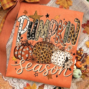 Może przedstawiać: Pomarańczowy t-shirt z napisem PUMPKIN SEASON pogrubioną czcionką. Wzór zawiera trzy dynie z różnymi motywami, w tym białymi kwiatami, panterką i czarnymi kropkami, ozdobione małymi gwiazdkami oraz liśćmi.