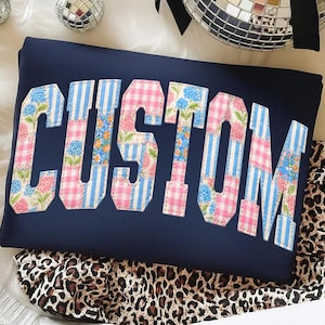 May include: Navy blue sweatshirt with the word "CUSTOM" in a patchwork design. The letters feature floral, gingham, and striped patterns in pink, blue, and white. The sweatshirt is resting on a leopard print fabric.