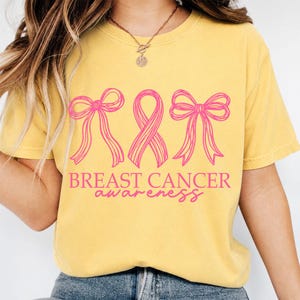 May include: A mustard yellow t-shirt featuring a pink graphic design of two bows flanking a central awareness ribbon. Below the graphic, the text reads BREAST CANCER awareness in a combination of block capital letters and a cursive script font.