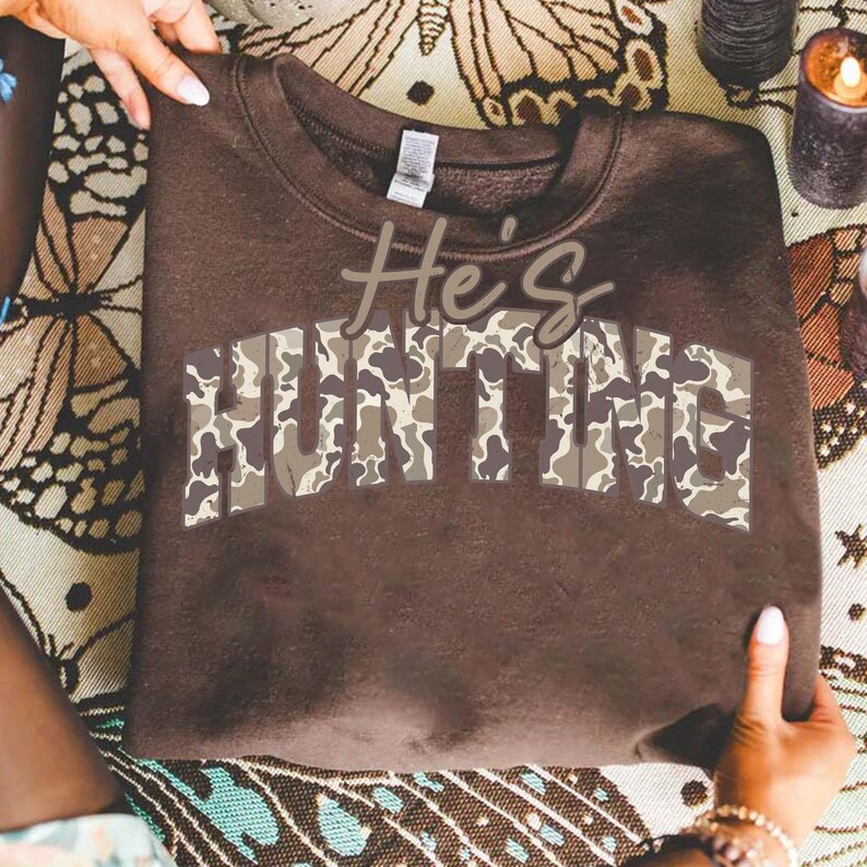 May include: A brown sweatshirt with the text "He's HUNTING" in a camouflage pattern. The sweatshirt is folded and held by hands, showcasing the design. The overall theme is related to outdoor activities and hunting.