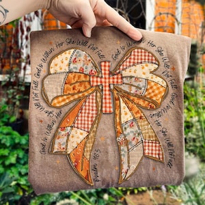 以下が含まれることがあります： 茶色の正方形の装飾品で、大きなリボンと十字架のデザインが特徴です。リボンはオレンジ、白、茶色のパッチワーク生地で作られています。「God Is Within Her She Will Not Fall」と「Psalm 46:5」の文字がプリントされています。