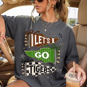 May include: A charcoal gray short-sleeve t-shirt featuring a graphic print of three pennants. The top pennant is brown with the text LET'S, the middle is green with GO, and the bottom is a black and white checkered pennant with TIGERS.