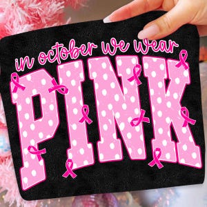 Puede incluir: Un parche de tela rectangular negro con el texto blanco "in october we wear" sobre las grandes letras rosas "PINK" con lunares blancos. Varias cintas rosas peque&ntilde;as de concienciaci&oacute;n sobre el c&aacute;ncer de mama est&aacute;n repartidas por las letras.