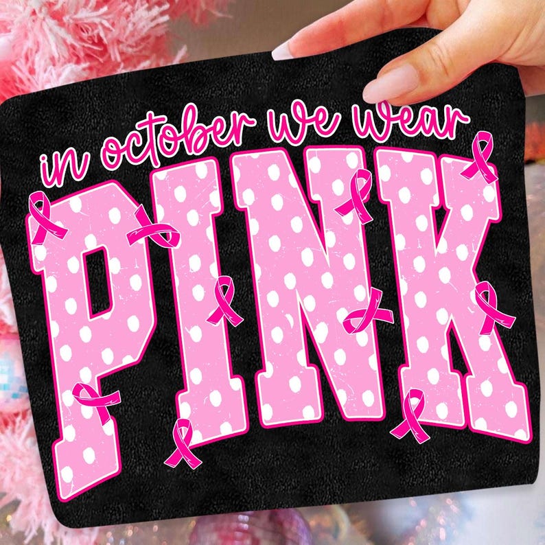 Puede incluir: Un parche de tela rectangular negro con el texto blanco "in october we wear" sobre las grandes letras rosas "PINK" con lunares blancos. Varias cintas rosas peque&ntilde;as de concienciaci&oacute;n sobre el c&aacute;ncer de mama est&aacute;n repartidas por las letras.