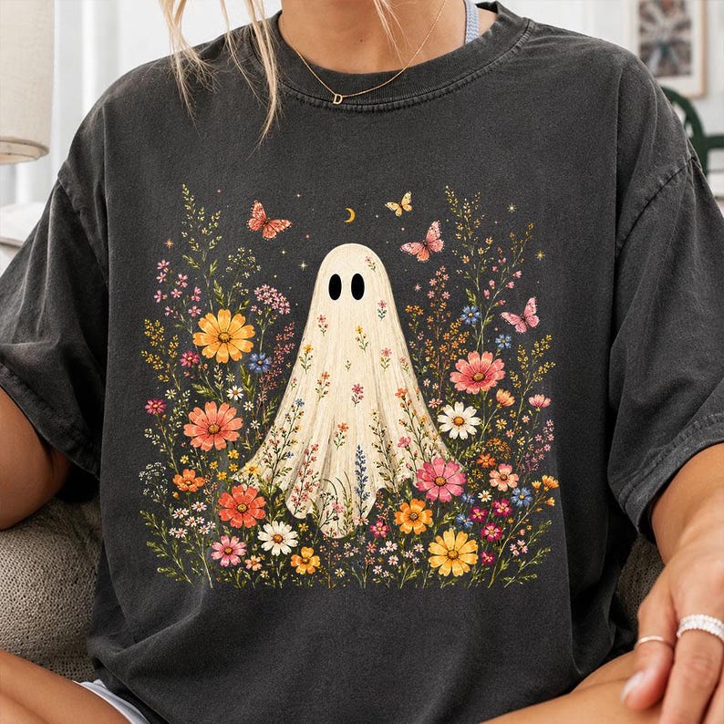 May include: A charcoal gray short-sleeve t-shirt featuring a central graphic of a white ghost surrounded by a vibrant array of colorful wildflowers, small butterflies, and a crescent moon. The design is detailed with various floral shapes.