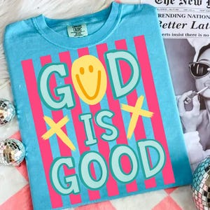 May include: A light blue t-shirt with the text "GOD IS GOOD" in a colorful design. The word "GOD" has a yellow smiley face in the center. Pink vertical stripes are behind the text.