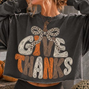 May include: A charcoal gray long-sleeve sweatshirt featuring a graphic of a pumpkin formed by the text GIVE THANKS in orange, white, and brown wood grain patterns, topped with a large leopard print bow. The garment has a raw, cropped hemline.