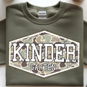 May include: Olive green sweatshirt with a crew neck. The front features a distressed, camouflage-patterned emblem with the word "KINDER" above "GARTEN" in white, block letters. The sweatshirt is made of heavy cotton.