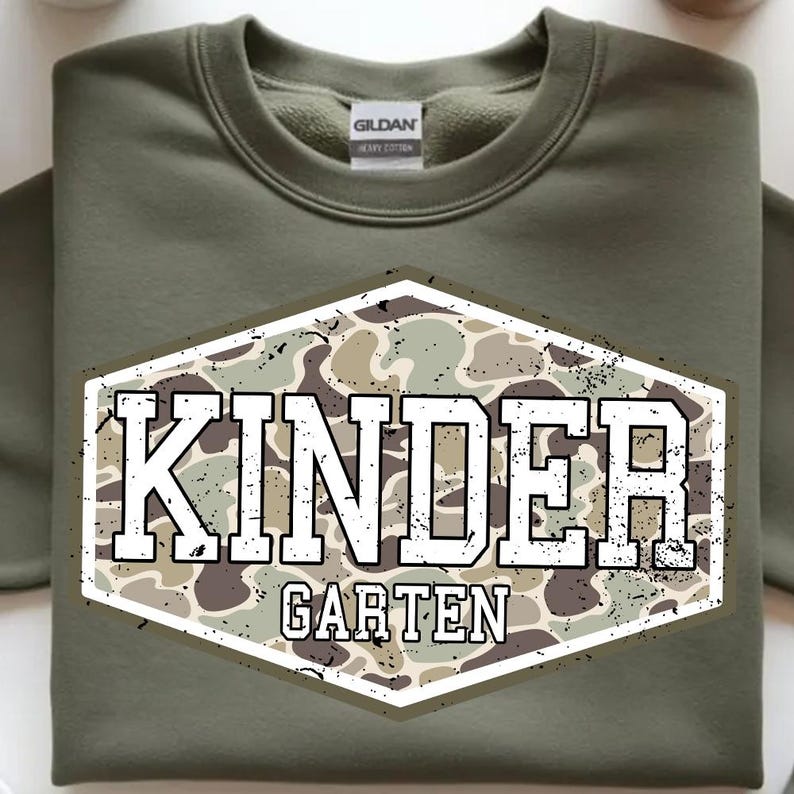 May include: Olive green sweatshirt with a crew neck. The front features a distressed, camouflage-patterned emblem with the word "KINDER" above "GARTEN" in white, block letters. The sweatshirt is made of heavy cotton.