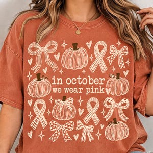 May include: A rust-colored t-shirt featuring a white graphic print of pumpkins, bows, and awareness ribbons. The text in the center reads, "in october we wear pink," surrounded by small heart and star icons. The design is arranged in a grid pattern.
