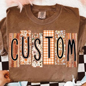 May include: A brown Comfort Colors t-shirt with the word "CUSTOM" in large, black outlined letters. Each letter is filled with a different fall-themed pattern, including florals and plaid.