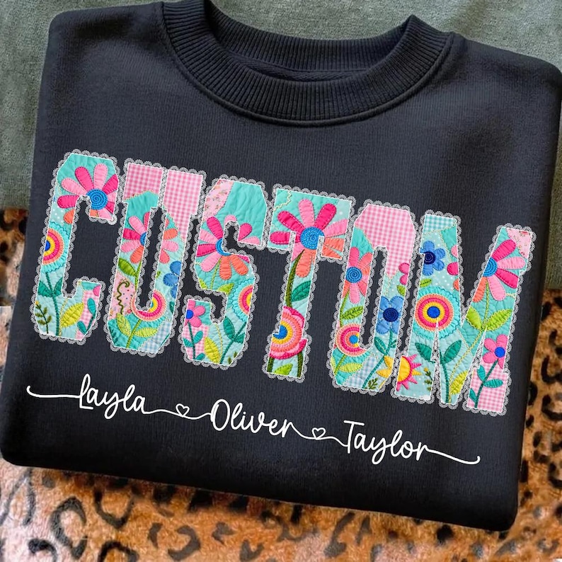 Op de afbeelding: Zwart sweatshirt met het woord "CUSTOM" in kleurrijke bloemenpatchwork letters. Daaronder staan de namen "Layla", "Oliver" en "Taylor" in witte letters, elk met een hartsymbool.