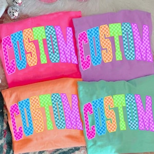 May include: Four colorful t-shirts in pink, orange, purple, and teal, each displaying the word "CUSTOM" in a checkered, rainbow-colored font. The shirts are arranged on a patterned surface, with a white cap and disco ball ornaments in the background.