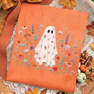 May include: An orange t-shirt featuring an embroidered white ghost character surrounded by a colorful field of wildflowers in shades of pink, blue, red, and purple. The ghost has two black oval eyes and is decorated with small floral patterns.