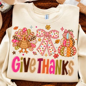 Puede incluir: Una sudadera de cuello redondo color crema con un estampado gráfico de un pavo, un lazo grande de lunares y una pila de tres calabazas, decorados con patrones coloridos de cuadros y lunares. El texto "GIVE THANKS" aparece en letras grandes y coloridas.