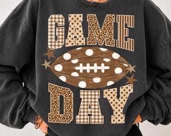 May include: A charcoal gray crewneck sweatshirt featuring the text GAME DAY in block letters with plaid, polka dot, and leopard print patterns. A brown football with white polka dots and white laces is centered between the words, accented by stars.