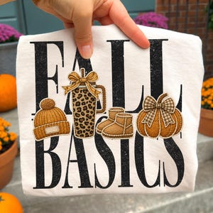May include: A white t-shirt featuring the black distressed text FALL BASICS. The design includes a mustard yellow knit beanie, a leopard print tumbler with a gold bow, tan suede-style boots, and an orange pumpkin with a brown gingham bow.