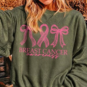 May include: An olive green long-sleeve sweatshirt featuring a pink graphic print of two bows flanking a breast cancer awareness ribbon. Below the graphic, the text reads "BREAST CANCER awareness" in a combination of block and script fonts.