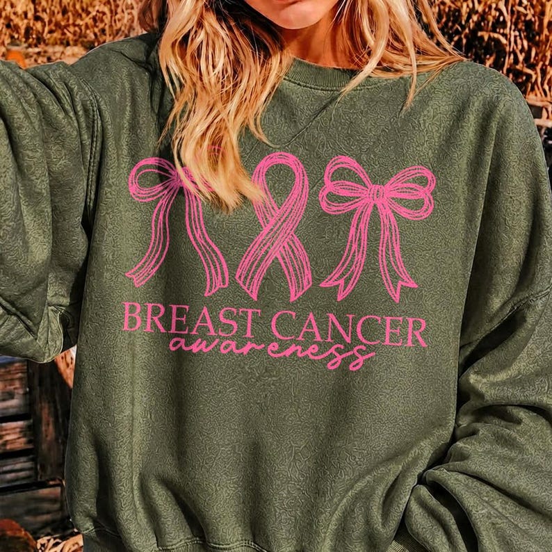 May include: An olive green long-sleeve sweatshirt featuring a pink graphic print of two bows flanking a breast cancer awareness ribbon. Below the graphic, the text reads "BREAST CANCER awareness" in a combination of block and script fonts.