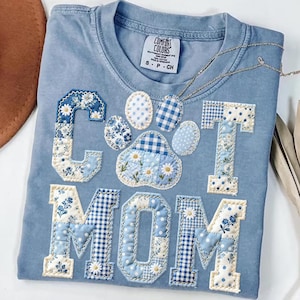Puede incluir: Una camiseta Comfort Colors azul claro con las palabras "Cat Mom" en un diseño de patchwork. Las letras y la huella de pata están hechas de varias telas florales y de cuadros azules y blancos. Un collar plateado está drapeado sobre la camisa.