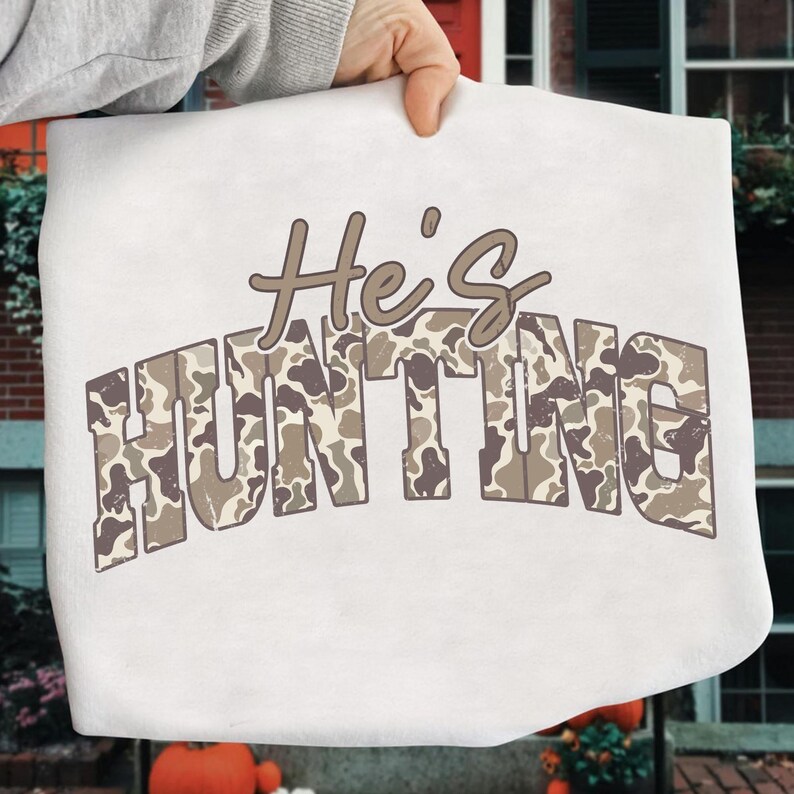 May include: A white sweatshirt with the text "He's HUNTING" in brown and camouflage print. The sweatshirt is held up by a person's hand. The text is in a bold, blocky font.