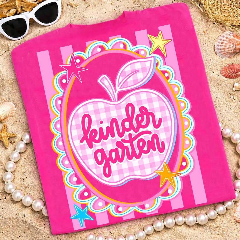 May include: A bright pink t-shirt with the words "Kindergarten" in a cursive font inside a gingham apple design. The shirt has vertical stripes and star accents. Sunglasses and a pearl necklace are in the background.