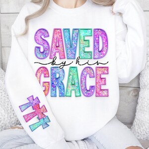 May include: White sweatshirt with the words "SAVED by his GRACE" in colorful, glittery letters. The sleeve features three crosses in matching colors. The design is a mix of pink, blue, green, and purple.