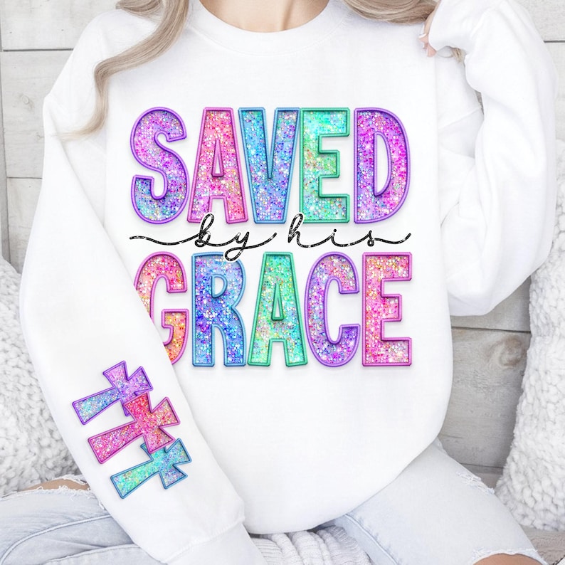 May include: White sweatshirt with the words "SAVED by his GRACE" in colorful, glittery letters. The sleeve features three crosses in matching colors. The design is a mix of pink, blue, green, and purple.