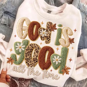May include: White sweatshirt with the embroidered phrase "God is Good all The Time." The text features a mix of textures, including leopard print, knitted, and floral designs, with autumn leaf accents. The sweatshirt is displayed on a denim jacket.