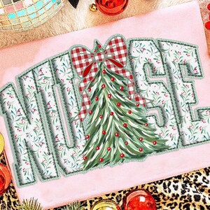 Op de afbeelding: Lichtroze t-shirt met het woord "NURSE" in een decoratief lettertype. De letters zijn wit met een groen bloemenpatroon en een groen gestikte rand. Een kerstboom en een rood geruite strik staan in het midden.