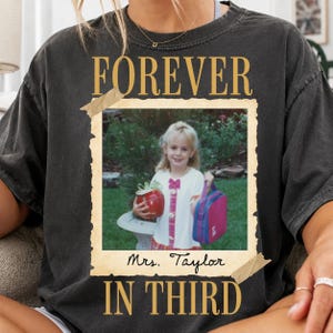 Op de afbeelding: Een antracietgrijs t-shirt met een vintage-stijl fotoprint van een jong kind dat een rode appel en een roze lunchtas vasthoudt. De tekst "FOREVER" staat boven de foto en "Mrs. Taylor IN THIRD" staat onder het fotokader.