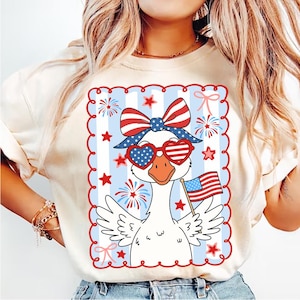 Puede incluir: Camiseta color crema con un dise&ntilde;o patri&oacute;tico de un ganso. El ganso lleva gafas de sol en forma de coraz&oacute;n y un lazo, sosteniendo una peque&ntilde;a bandera estadounidense. El dise&ntilde;o incluye elementos rojos, blancos y azules, estrellas y fuegos artificiales.