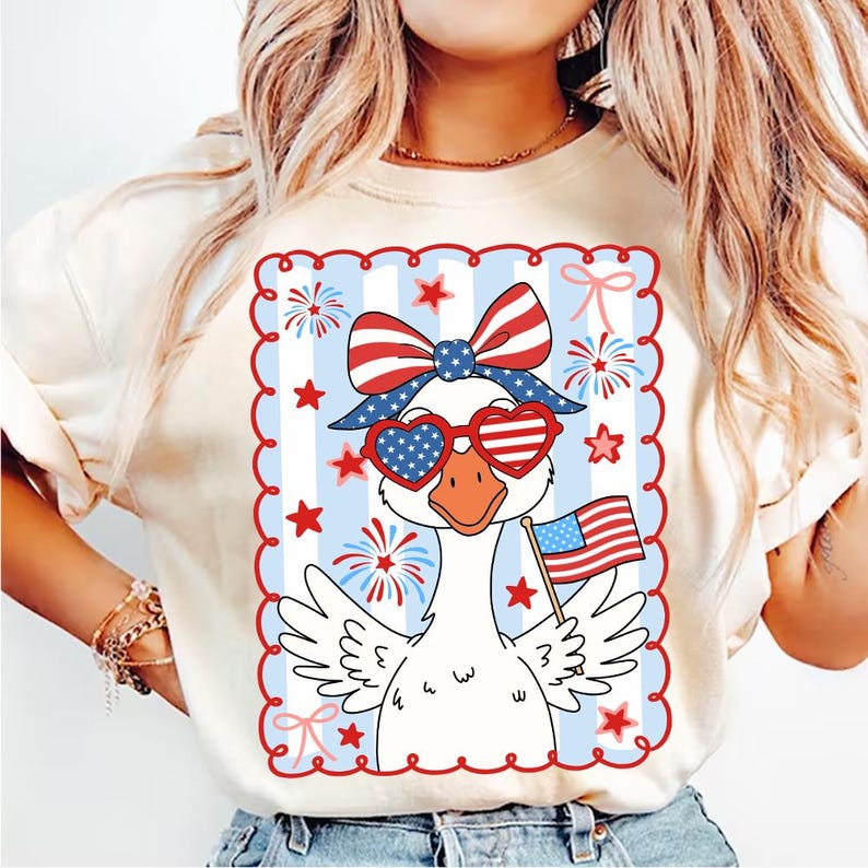 Puede incluir: Camiseta color crema con un dise&ntilde;o patri&oacute;tico de un ganso. El ganso lleva gafas de sol en forma de coraz&oacute;n y un lazo, sosteniendo una peque&ntilde;a bandera estadounidense. El dise&ntilde;o incluye elementos rojos, blancos y azules, estrellas y fuegos artificiales.