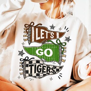 May include: A cream-colored crewneck sweatshirt featuring a graphic design of three pennants with the text LET'S GO TIGERS. The top pennant is brown, the middle is green with football field markings, and the bottom is a black and white checkerboard.