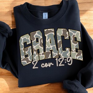 May include: Navy blue sweatshirt with the word "GRACE" in a camouflage pattern, outlined in gold. Below the word "GRACE" is the text "2 cor 12:9" in a script font. The sweatshirt is a Gildan brand.
