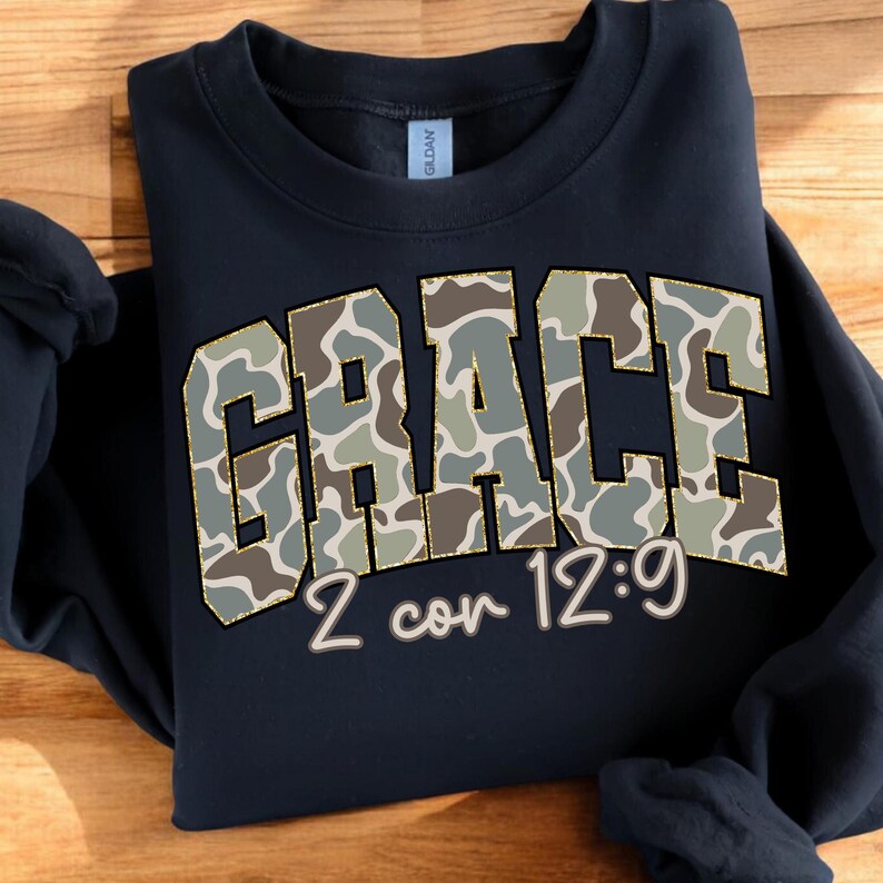 May include: Navy blue sweatshirt with the word "GRACE" in a camouflage pattern, outlined in gold. Below the word "GRACE" is the text "2 cor 12:9" in a script font. The sweatshirt is a Gildan brand.