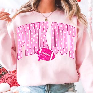 May include: A light pink crewneck sweatshirt featuring the text PINK OUT in large, bold, white-polka-dotted pink letters. Below the text is a pink football graphic with a breast cancer awareness ribbon integrated into the design.