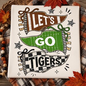 May include: A white square sign featuring three pennants with the text LET'S GO TIGERS. The top brown pennant says LET'S, the middle green football field pennant says GO, and the bottom checkered pennant says TIGERS, accented by small stars.