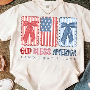 May include: White t-shirt with a patriotic design. The shirt features three rectangular frames with red, white, and blue elements, including bows and the American flag. The text "GOD BLESS AMERICA LAND THAT I LOVE" is printed below the frames.